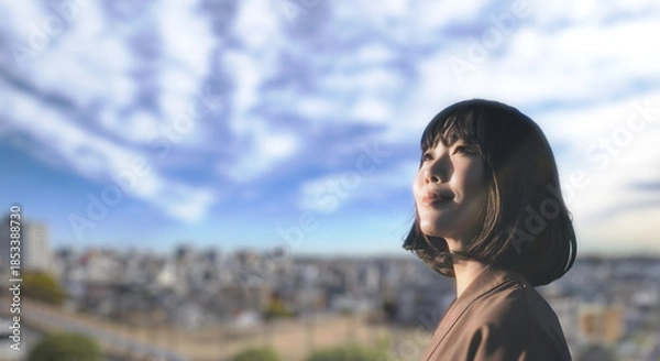 Obraz 上を見上げ微笑む明るい表情の日本人女性の顔　前向きでポジティブ希望に満ちた表情　就職・新生活・旅行・リラックス・入学のイメージ　青空と街の背景