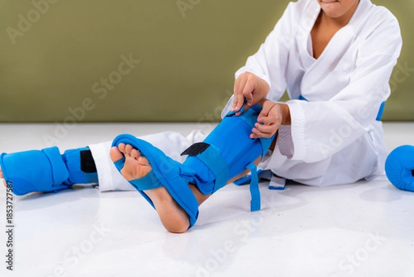 Obraz Boy wearing blue protective gear putting guards on legs before karate sparring match. Captures technique, safety focus, and organized preparation.