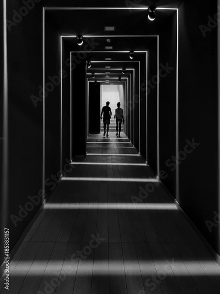 Obraz Silhouettes of two people in a corridor. Black and white image with bright light coming from behind and from few ambient lights. Couple walking away from the camera. Lens facing against the light.