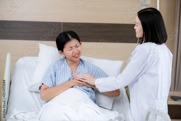 Fototapeta female patient lying in a hospital bed, appearing to be in pain as she clutches her chest with a pained expression. A doctor stands beside her, gently holding her hand and offering support while asses