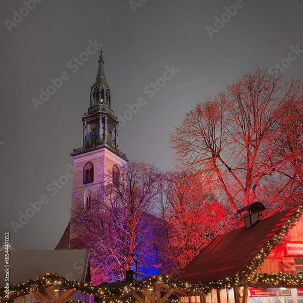 Obraz St. Mary’s Church tower rises behind colorful illuminated trees and festive wooden stalls at the Christmas market on Alexanderplatz in Berlin on a winter night.