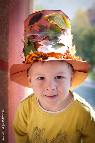 Fototapeta A child stands outside a house wearing a colorful hat made of leaves and flowers. The sun is shining, and the leaves show bright autumn colors.
