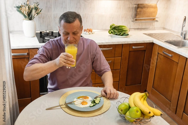 Obraz Mature man drinking orange juice and having a healthy homemade breakfast in kitchen.