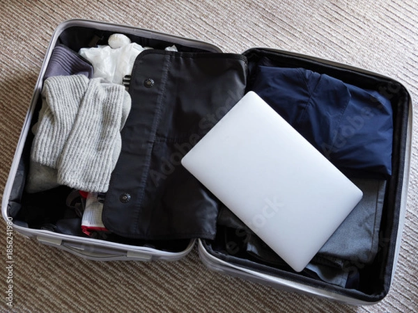 Obraz 家のリビングの絨毯の上にある出張用のスーツケースと中身の衣服類とノートパソコンの様子