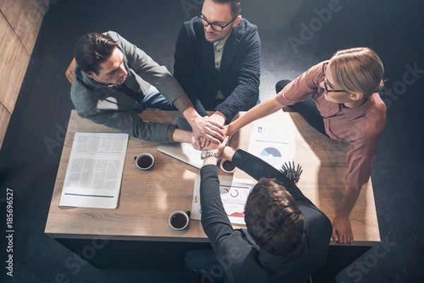 Fototapeta Top view happy female and glad men putting hands on each other while sitting at desk in apartment. Labor concept