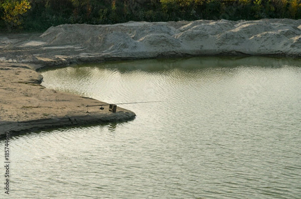 Fototapeta Fishing from the cape of the sandy coast. Sand dunes surround the lush vegetation. Vague shadows of trees and coast. Small waves on the surface disrupt the peace. 