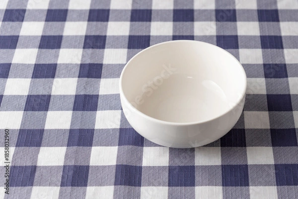 Fototapeta Empty white ceramic bowl on a blue and white checkered tablecloth with copy space on the left. Minimalist tableware for cereal or soup