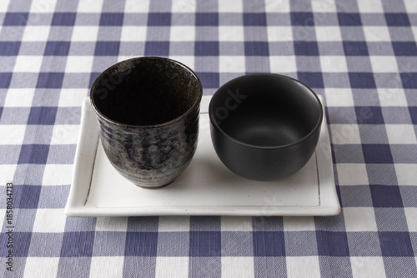Fototapeta Two different black ceramic cups on white rectangular plate over blue checkered tablecloth