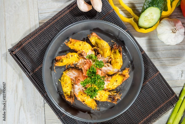 Obraz A round black plate with grilled shrimp arranged in a circle, garnished with parsley, surrounded by fresh vegetables on a wooden mat.