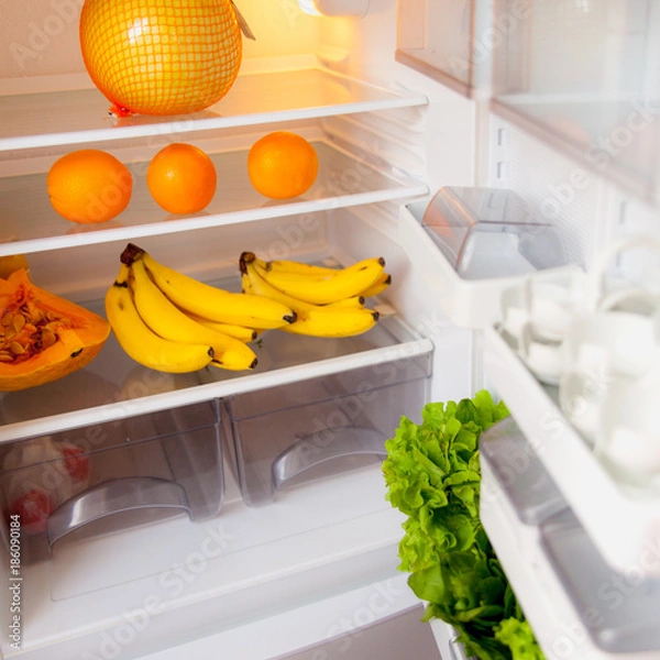 Fototapeta Open fridge with fruit and greens inside on white shelves. The concept of a healthy diet.