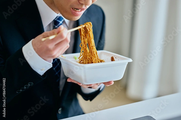 Fototapeta カップ焼きそばを食べながら仕事をする20代の若い男性
