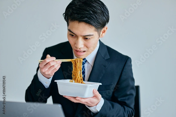 Fototapeta カップ焼きそばを食べながら仕事をする20代の若い男性