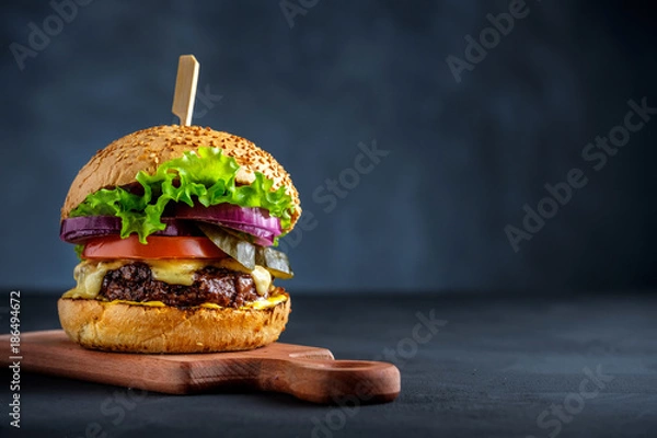 Fototapeta Smaczny grillowany burger wołowy z sałatą, serem i cebulą podawany na desce do krojenia na czarnym drewnianym stole, z copyspace.