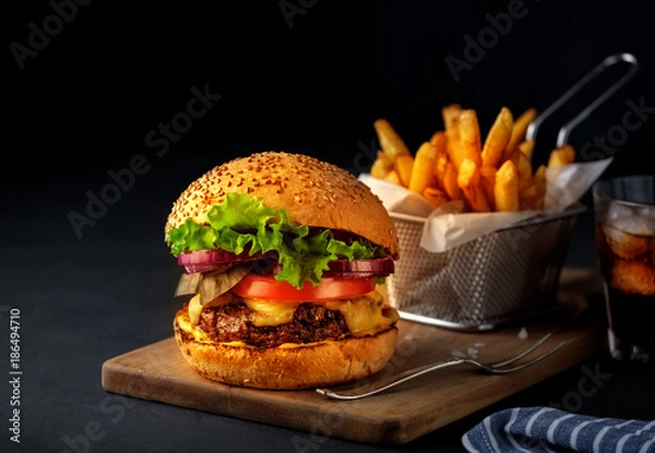 Fototapeta Tasty grilled beef burger with lettuce, cheese and onion served on cutting board on a black wooden table, with copyspace.