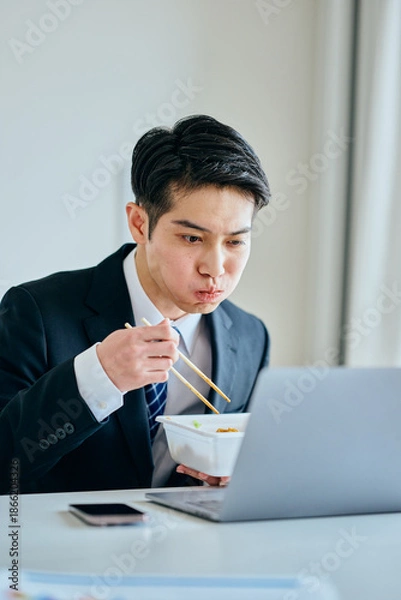 Fototapeta カップ焼きそばを食べながら仕事をする20代の若い男性