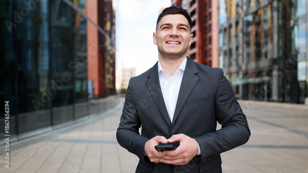 Fototapeta Man stands outside buildings while using smartphone