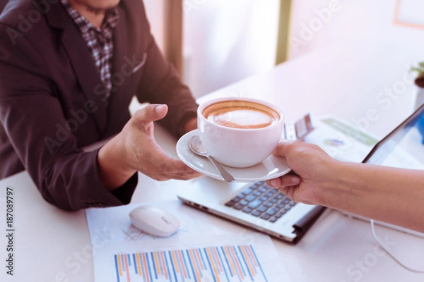 Fototapeta Close up of man's hand taking coffee cup from woman's hand in coffee shop. Portrait of hand holding white cup of cappuccino in office or co-workspace.