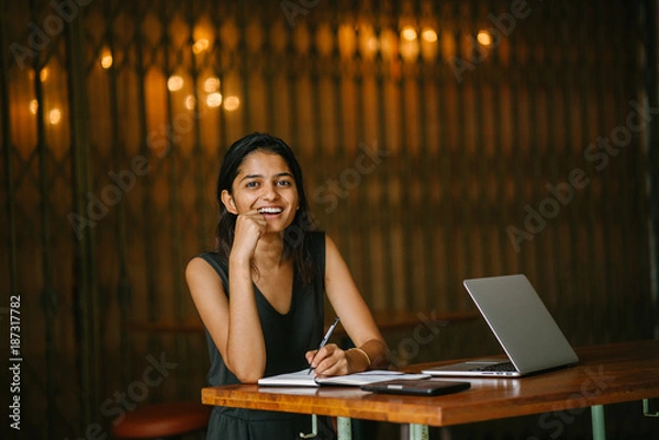 Fototapeta Portrait of Indian lady writing in a notebook in a cosy cafe with a laptop nearby