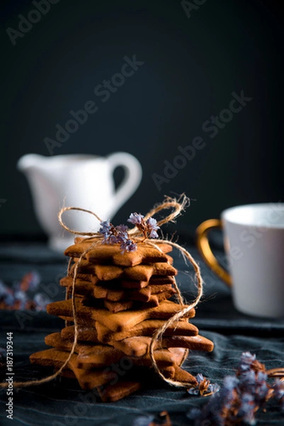 Obraz Tied stack of star shaped gingerbread cookies, white cup with a gold handle and a white ceramic milk pot. Decorated with dried violet flowers, dark background.