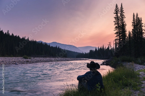 Obraz Cowboy sitting by a raging river at sunset. The gorgeous colors of an Alberta sunset are displayed as the green grass grows and the river rushes by. The man sits with a denim jacket and cowboy hat.