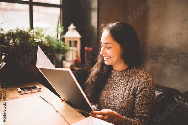 Fototapeta business girl studying the menu in restaurant decorated with Christmas decor.sits near the window on cloudy winter day at wooden table.Dressed in warm gray sweater.On the table,phone and glasses