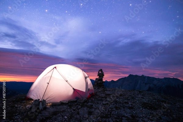 Obraz Lit up tent under the stars on a mountain. There is light pollution that looks like a sunset or sunrise with a layer of clouds in between the starry night sky. There is an inukshuk beside the tent.
