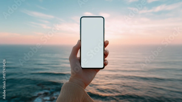 Obraz 穏やかな海と空が広がる夕暮れ時（または朝焼け）の浜辺で、白い画面のスマートフォンを水平線に向けて掲げる手元、モックアップ