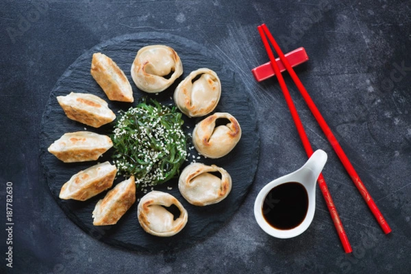 Fototapeta Pan fried asian dumplings served on a stone slate with chuka salad and dipping sauce. Flat-lay on a dark scratched metal background, horizontal shot