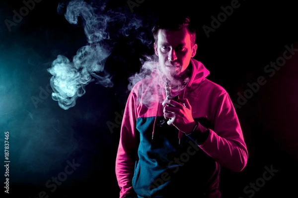 Obraz The man smoke an electronic cigarette  on a background of red and blue smoke