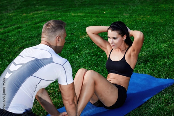 Obraz The coach on the grass holds the legs of the girl and helps exercise the abdominal muscles.