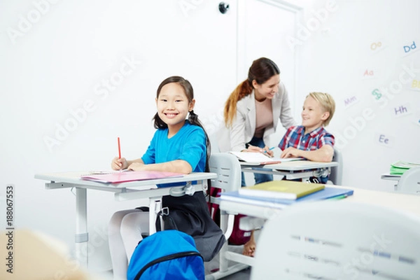 Fototapeta Happy schoolgirl looking at camera by desk with her teacher and classmate talking on background