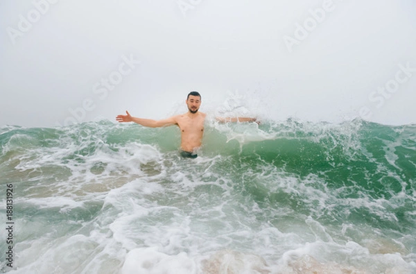 Obraz Odd bizarre crazy fearless awesome adult man enjoying in cold stormy sea. Guy jumping in waves in ocean with emotional funny face. Adult male fooling like child in water outdoors. Leisure activities