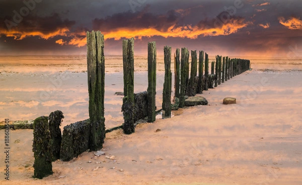 Fototapeta fiery sunset sky over old water breaker, groyne on a wet sandy beach