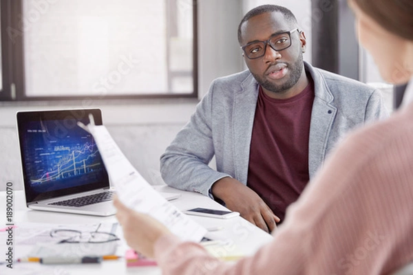 Fototapeta Photo of serious dark skinned male manager and female financier collaborate together, anticipate income, do paper work, use laptop computer for creating graphics to illustrate company financial status