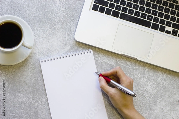 Fototapeta Modern office table desk, flat lay, top view. Workplace with a laptop, coffee cup, woman is redy to write in a blank notepad, grey concrete background
