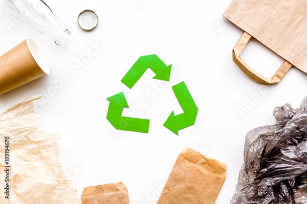 Obraz Sorting waste and recycle. Green paper recycling sign among waste paper, plastic, glass, polyethylene on white background top view