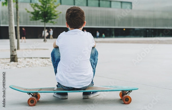 Fototapeta Back view. The boy, dressed in a white T-shirt and blue jeans, sits on a city street on a skateboard. On background modern building in soft focus. Lifestyle. Space for text, logo, image. Mock up.