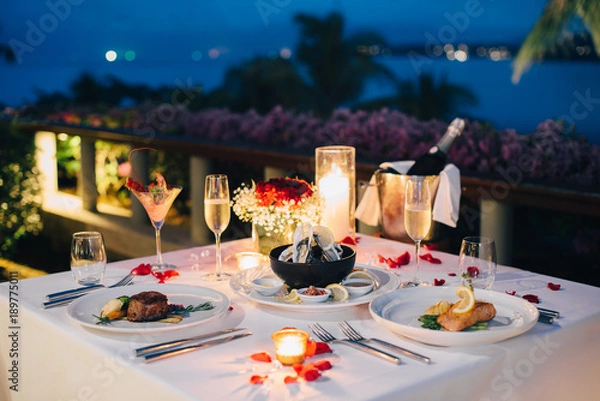Fototapeta Romantic candlelight dinner luxury table setup for couple with beautiful light as background. Glasses of champagne and beautiful food presentation on table.. Concept for valentine's day  and date.