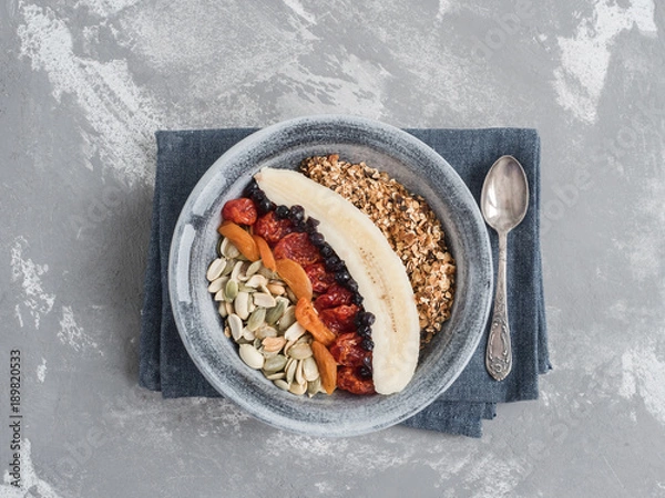 Fototapeta Muesli with banana, blueberries, dried apricots, carnelian cherry, peanuts and pumpkin seeds, in an old plate on a gray table