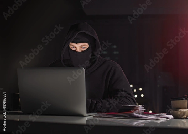 Obraz Portrait of orderly hacker typing in appliance while sitting at table in dark apartment. Crime and digital device concept