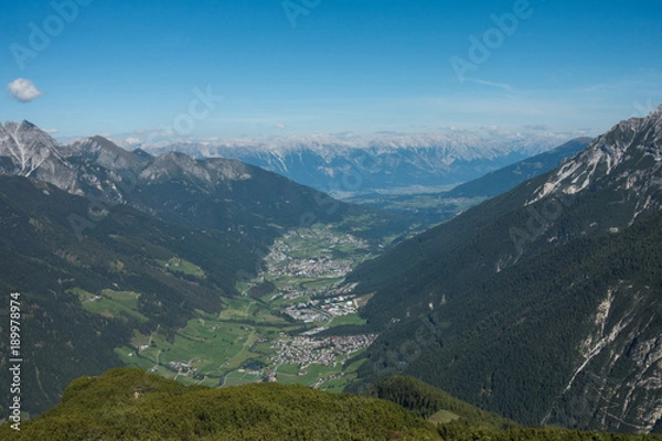 Fototapeta View overlooking a town in a valley between two mountain ranges. There is a small town in the valley with green space all around and forest leading up the sides of the mountains.