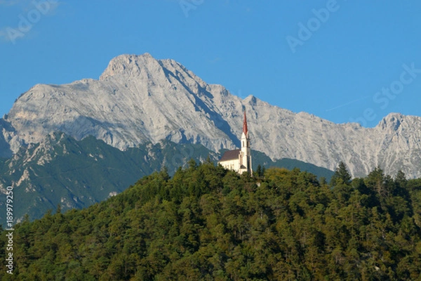 Obraz Church on a hill in a valley of the Austrian Alps The church sits among trees high above a roadway. The church has orange tile roofing and contrasts the greenery nicely.