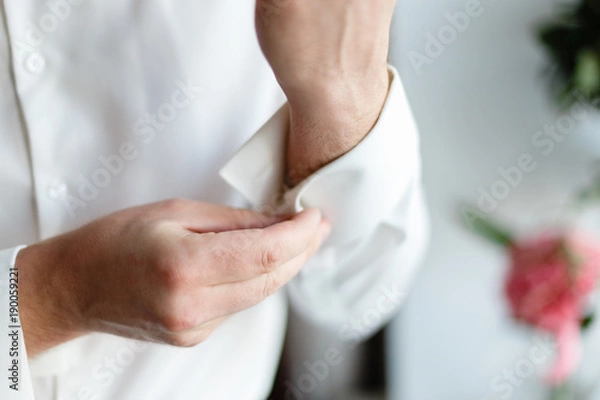 Fototapeta A man wears buttons of cufflinks on the sleeves of the white shirt. Businessman getting dressed by the window.