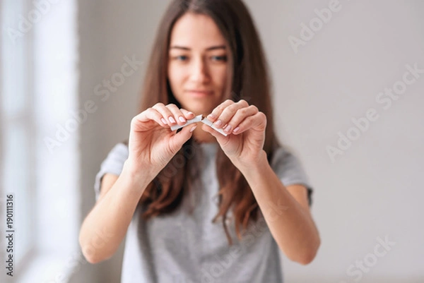 Fototapeta Stop smoking cigarettes concept. Portrait of beautiful smiling girl holding broken cigarette in hands. Happy female quitting smoking cigarettes. Quit bad habit, health care concept. No smoking.