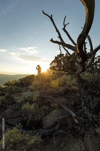 Fototapeta A cowboy tips his hat on top of a mountain as golden rays from the sun shine behind him.