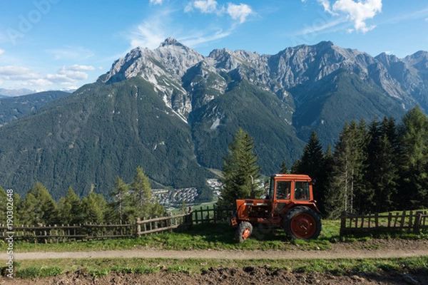 Obraz An orange tractor sits on a hill overlooking a town in the Alps. There is an old path and a fence beside the tractor. There are trees behind the tractor and fence line.