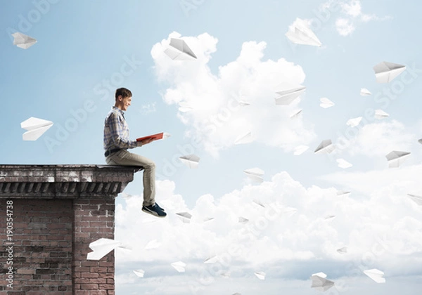 Fototapeta Handsome student guy on roof edge reading book and paper planes fly around