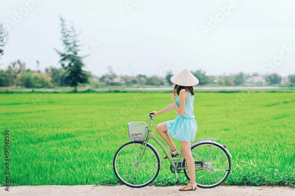 Fototapeta A girl on a bicycle in a traditional Vietnamese hat stands on the road and admires the rice fields.The girl rides a bicycle in a traditional Vietnamese hat in the midst of rice fields, Hoi An, Vietnam