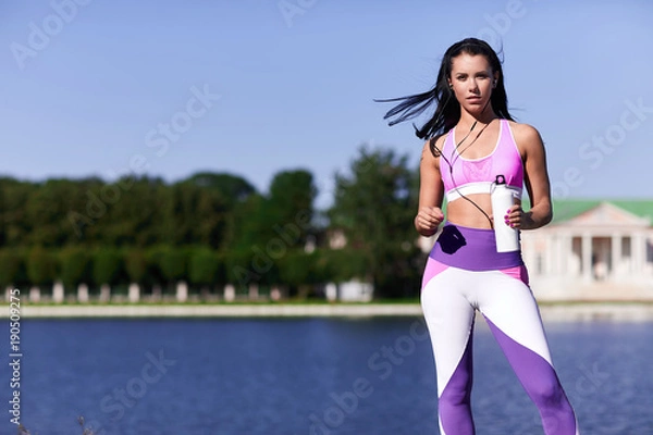 Obraz Female athlete standing near the river with a bottle of water and looking forward.