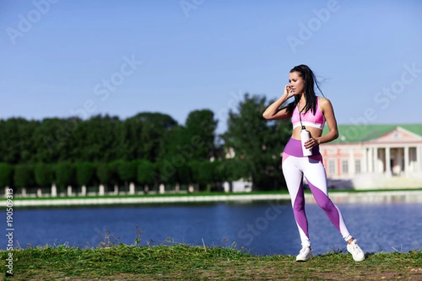 Obraz Woman in sports clothes standing on the riverbank with a bottle of water and looking to the left.
